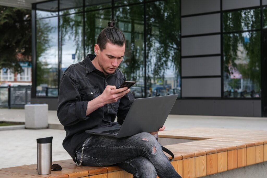 A young guy sends a text message to a mobile phone and works on a laptop